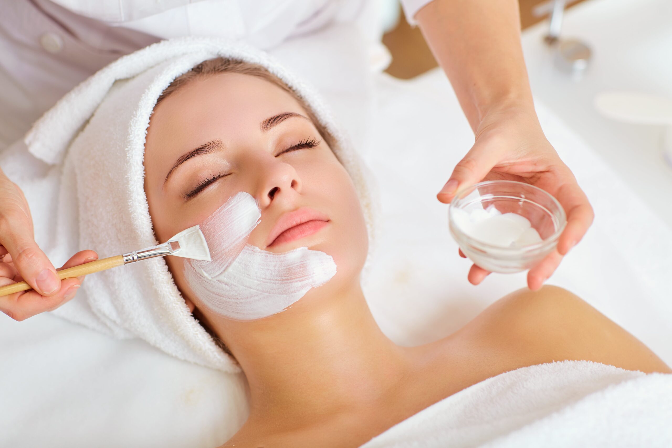 A young woman with a towel wrapped around her head receives a relaxing facial treatment. A spa professional is applying a white facial mask with a brush while holding a small glass bowl of cream. The woman appears calm and serene.